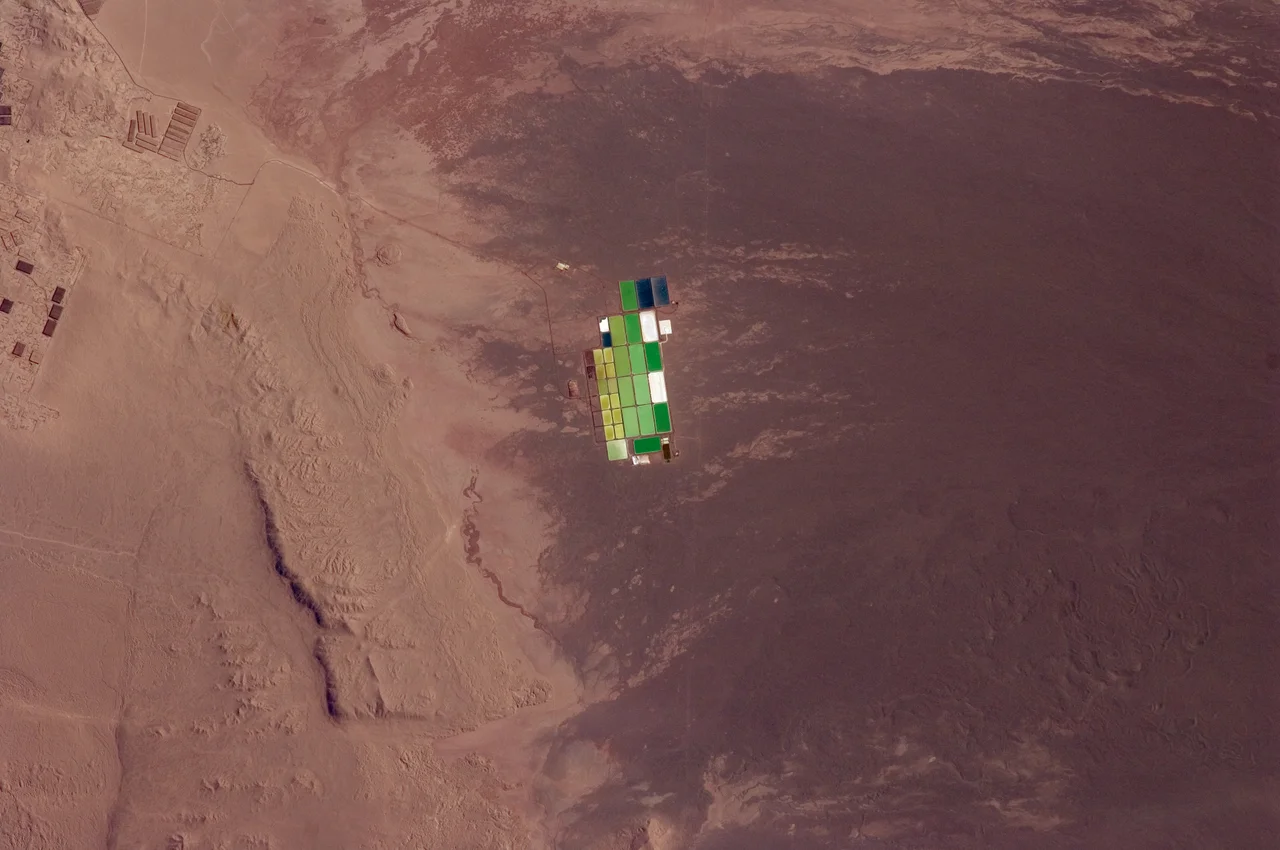Brightly coloured solar evaporation (salt) ponds in a desert landscape give this astronaut photo an unreal quality. The ponds sit near the foot of a long alluvial fan in the Pampa del Tamarugal, the great hyper-arid inner valley of Chile’s Atacama Desert. The alluvial fan sediments are dark brown, and they contrast sharply with tan sediments of the Pampa del Tamarugal. Nitrates and many other minerals are mined in this region. A few extraction pits and ore dumps are visible at upper left. Iodine is one of the products from mining; it is first extracted by heap leaching. Waste liquids from the iodine plants are dried in the tan and brightly coloured evaporation ponds to crystallize nitrate salts for collection. Coordinates: -20.946162, -69.514961
This image was aquired with a Nikon D2X digital camera fitted with a 400 mm lens, and is provided by the ISS Crew Earth Observations experiment and Image Science & Analysis Laboratory, Johnson Space Centre.