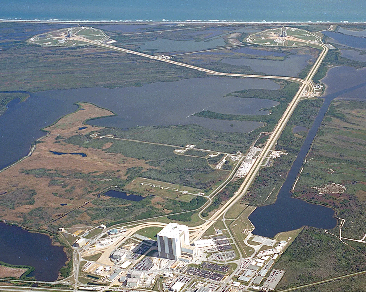 KSC's Launch Complex 39 is strategically located next to a barge site and a variety of structures, including a Vehicle Assembly Building (VAB), Orbiter Processing Facilities (OPF), Press Site, Launch Control Center (LCC), and a crawlerway to the pads.
The crawlerway, leading to pads 39B on the left and 39A on the right, can be seen extending from the massive VAB at the left in this photo. The VAB, which covers eight acres and stands 525 feet tall, is used for assembly, stacking and mating of Space Shuttle elements. Originally built for assembly of Apollo/Saturn vehicles and later modified to support Space Shuttle operations, the VAB is one of the largest buildings in the world.
The LCC, seen here as the small white building to the upper right of the VAB, is where launch, mission support, and loading are controlled.