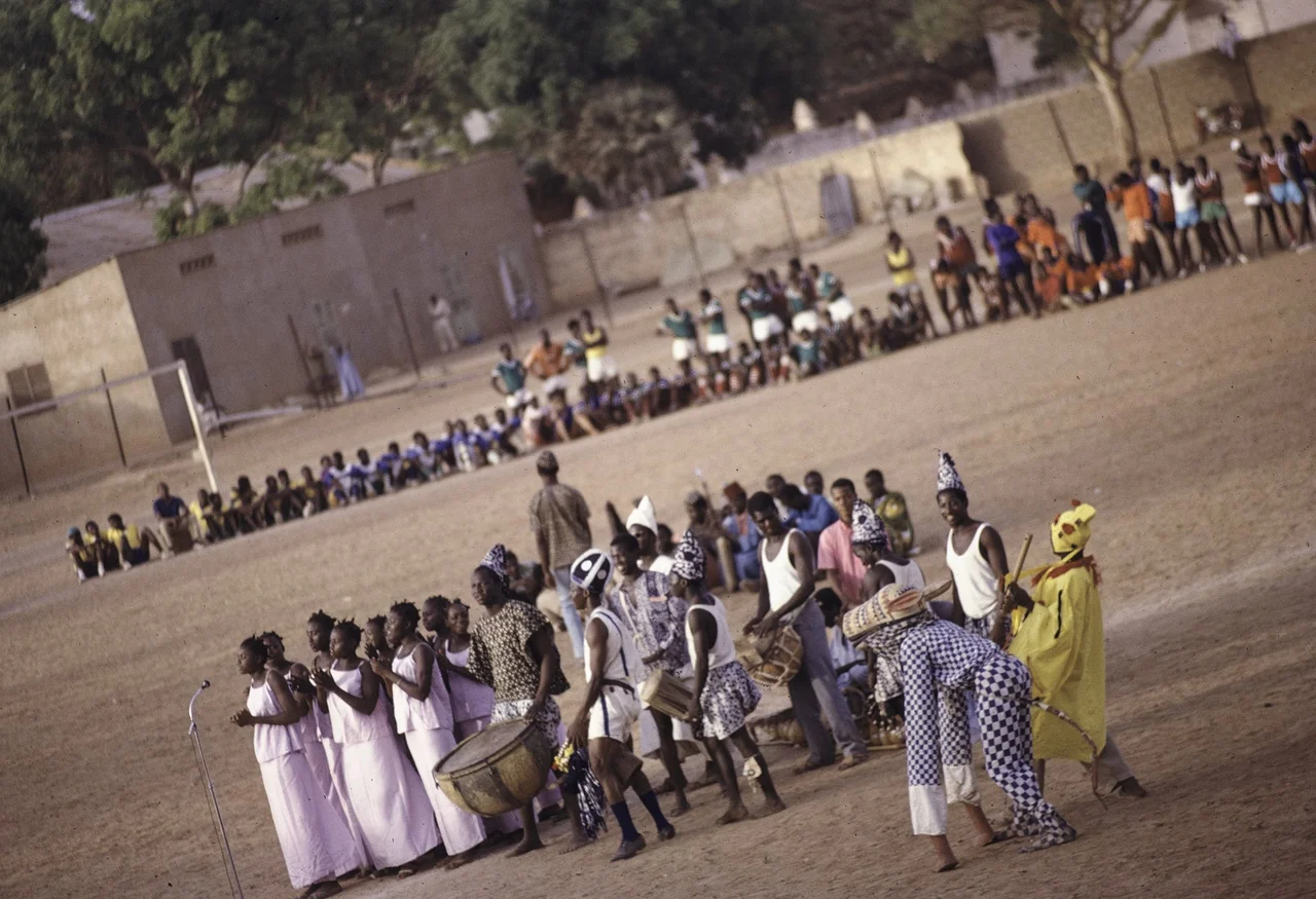 The group represents Ségou. Eight traditional female singers clad in white are standing behind a microphone. A man is carrying a hugekettle drum, three other men with tall caps are handling smaller drums. Two masked and disguised dancers, one as a donkey in checkered clothes. A calabash drum, a balafon, a long flute wielded by a man in yellow. A line of spectators at the edge of the football field.