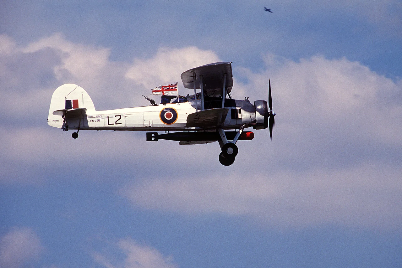 A Royal Navy Fairey Swordfish Mk. II (serial number LS326) in flight during Air Fete'88, a NATO aircraft display hosted by the U.S. Air Force's 513th Airborne Command and Control Wing, at RAF Mildenhall, Suffolk (UK). This aircraft is today operated by the Royal Navy Historic Flight.
RN description: This aircraft was built in 1943 at Sherburn-in-Elmet. Later that year she was part of 'L' Flight of 836 Squadron on board the MAC ship Rapana, on North Atlantic Convoy duties. Following her active service she was used for training and communications duties from the Royal Naval Air Station Culham near Oxford and Worthy Down near Winchester. In 1947 Fairey Aviation bought LS326 and displayed her at various RAeS Garden party displays. The following year she was sent to White Waltham for storage and remained there getting more and more dilapidated until Sir Richard Fairey gave orders for the aircraft to be rebuilt. The restoration work completed in October 1955 and thereafter she was kept in flying condition at White Waltham registered as G-AJVH and painted Fairey Blue and silver. In 1959 LS326 was repainted for a starring role in the film 'Sink the Bismarck!'. In October 1960 she was presented to the Royal Navy by the Westland Aircraft Company and has been flown ever since. For many years she retained her "Bismarck" colour scheme and in 1984 D-Day invasion stripes were also added for the 40th Anniversary celebrations when she overflew the beaches of Normandy. Since 1987 she has worn her original wartime colour scheme for North Atlantic convoys with 'L' Flight of 836 Squadron. Following extensive work by BAeS Brough to her wings, it is hoped to have LS326 back in flying condition again in 2008.