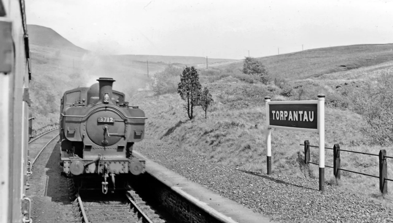 Torpantau Station: meeting a Brecon - Newport train.
View northward, towards Torpantau Tunnel, Talybont and Brecon: ex-Brecon &amp; Merthyr (Brecon - Dowlais section), later GWR. The down train, headed by standard '8750' class 0-6-0PT No. 3712 (built 11/36) has just surmounted the fearsome climb of seven miles (the "Seven-Mile Bank") at about 1-in-40 from Talybont-on-Usk, while my train headed for Brecon has come up a similar climb from Pontsticill Junction. The narrow-gauge Brecon Mountain Railway from Pant terminates just short of here but aims to reach this point. On the left is the outline of the Brecon Beacons.