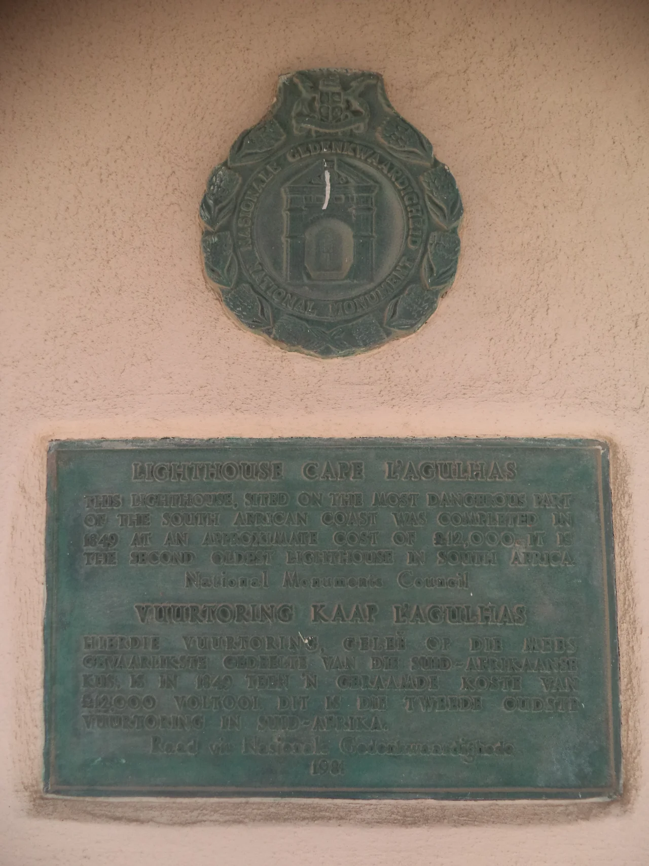 The memorial seal and the memorial plaque on the entrance of Cape Agulhas Lighthouse. The inscription on the seal writes:
National Monument
The inscription on the plaque writes:
Cape Agulhas Lighthouse
This lighthouse, sited on the most dangerous part
of the South African coast was completed in
1849 at an approximate cost of £12,000. It is
the second oldest lighthouse in South Africa.
National Monument Council