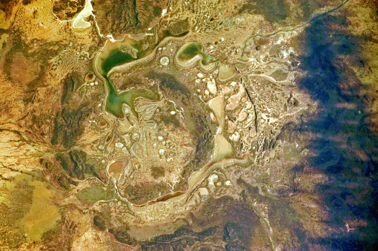 The Shoemaker impact site is approximately 30 kilometres in diameter and clearly defined by concentric ring structures formed in sedimentary rocks (brown to dark brown, image centre).
Several saline and ephemeral lakes—Nabberu, Teague, Shoemaker, and numerous smaller ponds—occupy the land surface between the ring structures. Differences in colour result from both water depth and from suspended sediments, with some bright salt crusts visible around the edges of smaller ponds (image centre).