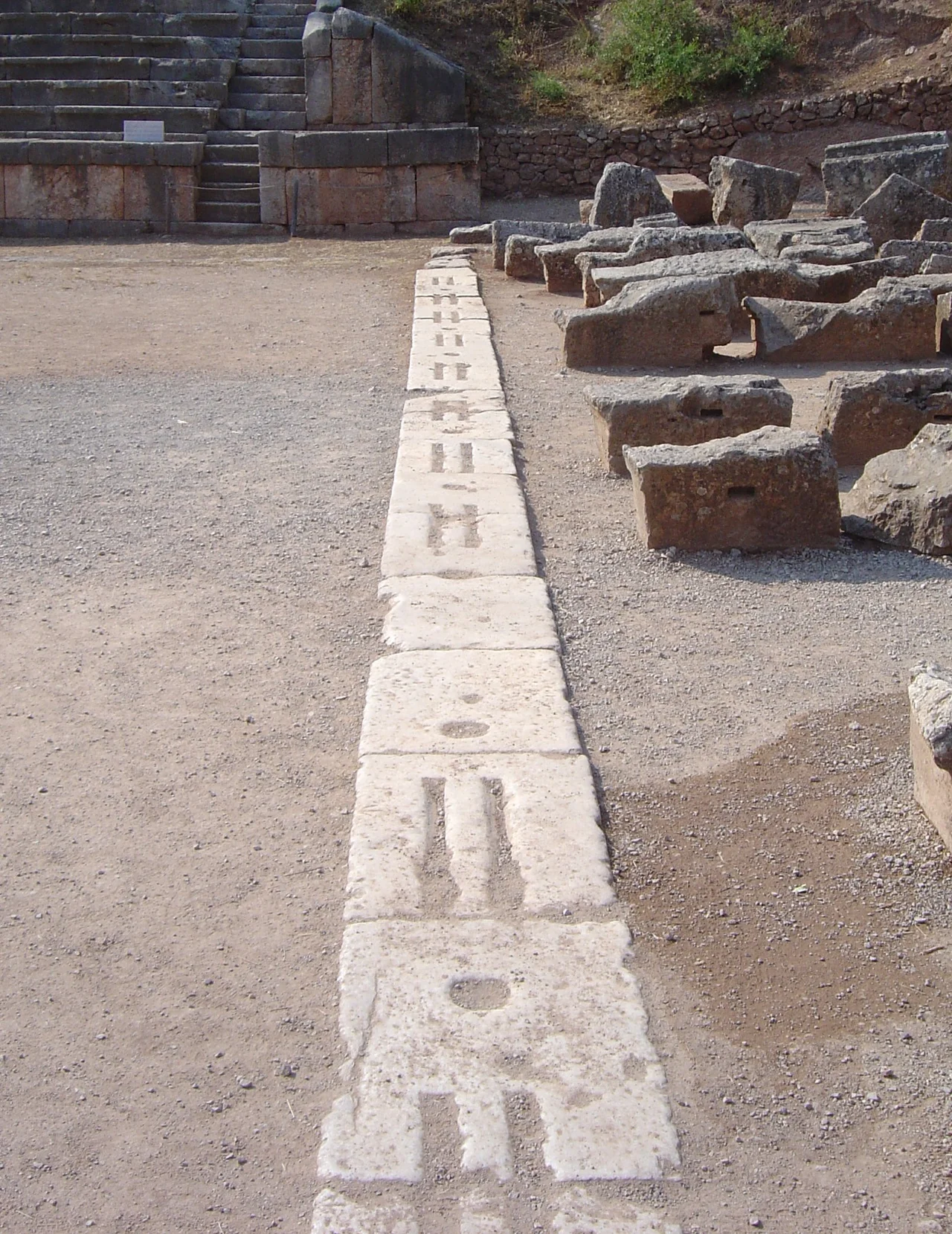 The starting line at the stadium used for the Pythian Games at Delphi, Greece. This starting line has a design representative of that of many ancient Greek stadiums: stones with two lines in which the athletes nudged their toes, and round holes in which posts could be erected to support the start signalling mechanism.
The stone steps for sitting the public behind were erected under the Romans.