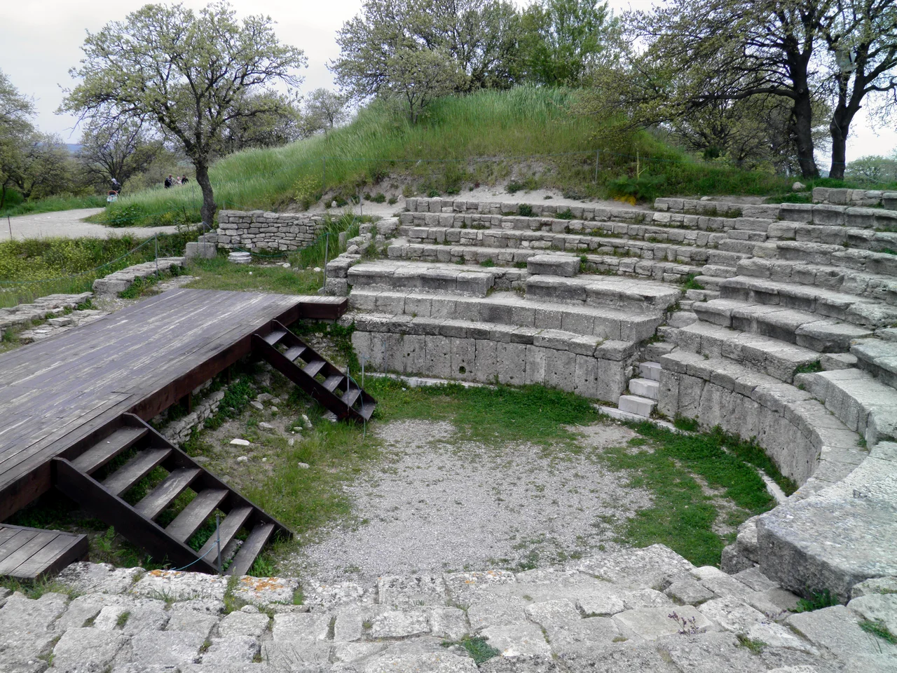 Hadrianic Odeon in Troy IX, small Theatre renovated by Hadrian in 124 AD.
In the early second century AD the provincial city of Ilion built a new theater unlike any architecture it had previously realized. At that time Roman Ilion was still best known for the large Hellenistic sanctuary of Athena (begun ca. 230 BC) with its imposing white marble temple complex bedecking the city’s acropolis. In contrast, the new theater was very small (perhaps the smallest in the whole Aegean area) and had an opulent scenae frons, or stage building. It was first discovered by Heinrich Schliemann’s architect and successor, Wilhelm Dörpfeld, in 1893, but received scant attention from archaeologists at Troy until recently (Aylward 2000, p. 138). Because of its diminutive size the current archaeologists from the University of Cincinnati name it an “odeion,” place for recitation of odes.
Briefly described, the Odeion of Ilion had a two-storey scenae of very limited depth, even including the aediculated bays. The foundation tells us how limited in depth the whole construction was, and it also shows an irregular column spacing mirrored about the center. The center opening is slightly wider than the pairs of openings to either side, and the pairs of columns which frame each opening are spaced even closer together. This inflected spacing of the vertical elements tells us the projecting and receding sections of entablature repeat in the upper storey in alignment (or stacking) with the lower entablature. This is in noticeable contrast to the trend since the late first century BC in Miletus and Ephesos where the spacing of the vertical elements is uninflected, thus allowing the projecting entablatures to alternate (or offset) from storey to storey without gross distortion of proportions. A secondary effect of the “in alignment” type of scheme is a greater inflection toward and emphasis of the center; at Ilion the Odeion has a large broken pediment spanning the center three bays at the upper storey, and at the lower storey entablature level there is no concavity, as expected by the normal rules. The upper storey also confounds the rules by including three different orders: Ionic for the major columns, Pergamene for the center bay, and Corinthian for the secondary niches. The whole scheme was a carefully orchestrated composition in white marble elements juxtaposed to colored marbles (blue-grey, violet, pink, coral, red, beige, green and yellow).
The emperor Hadrian visited the city in 124. He appears to have ordered new repairs and may have redecorated the odeon, where his statue has been found. His visit marks the beginning of a golden age, with the construction of Roman baths, a fountain (nymphaeum), and an aqueduct. Substantial parts of it survive and can be seen near Kemerdere.