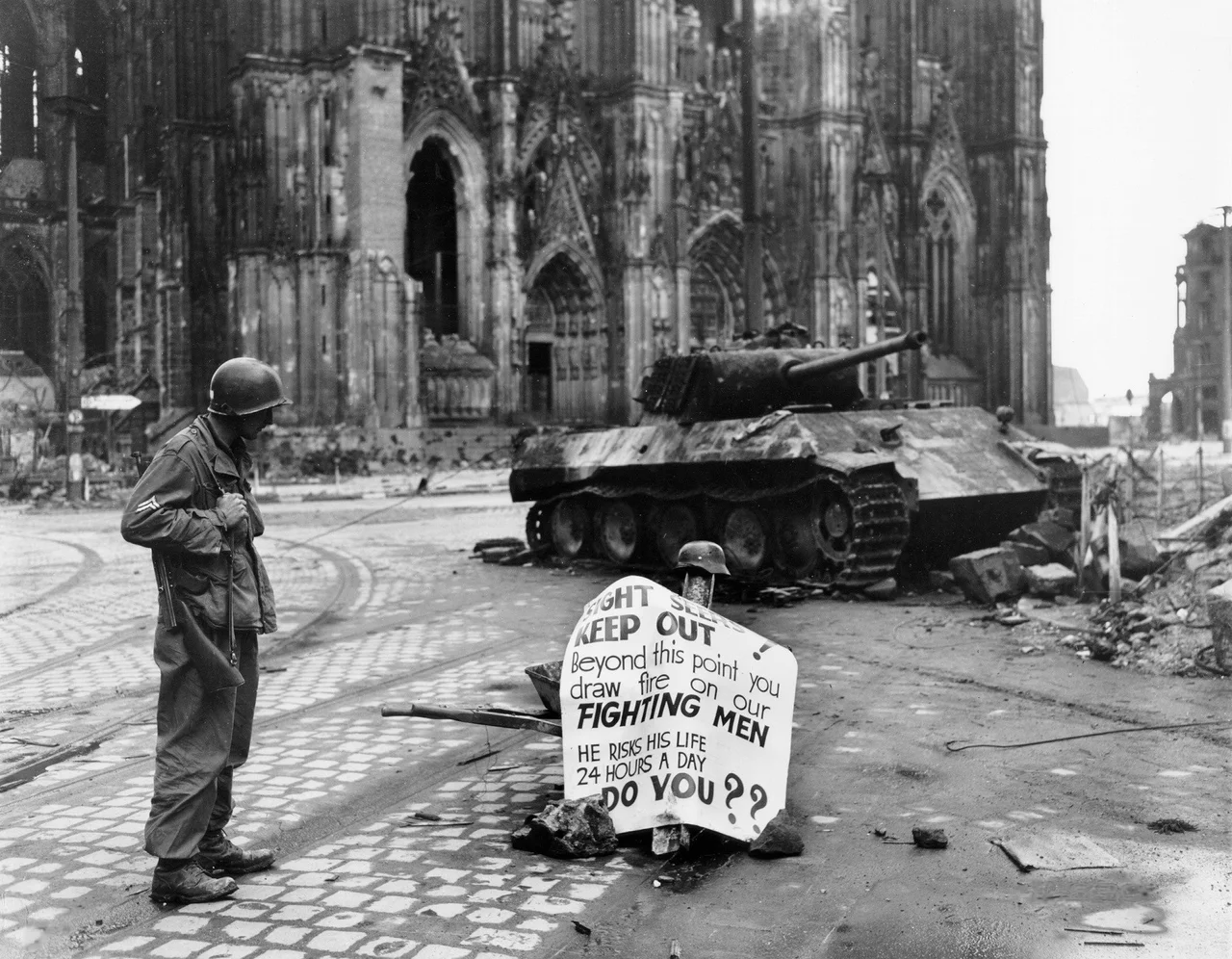 Original caption: Cologne, Germany - Cpl. Luther E. Boger, Concord, N.C., skytrooper, reads a warning sign in the street. This street leads to the Rhine River and is under observation of the Germans who occupy a stronghold there. Cpl. Boger is with the 82nd Airborne Division. 4 April 1945. The German tank is burnt out and the torsion-bars have been destroyed by the immense heat of the fire.
Allied troops captured the western part of Cologne on 6th-7th March 1945. The German army still held the eastern shore of the Rhine and attacked the Allies with artillery. The rest of Cologne was captured between 12th and 15th April 1945. Until the 16th April a strip of about 500m along the shore had been declared as a restricted area and the cathedral was just within this zone.
Text on sign reads:
SIGHT SEERS
KEEP OUT!
Beyond this point you
draw fire on our
FIGHTING MEN
HE RISKS HIS LIFE
24 HOURS A DAY
DO YOU??