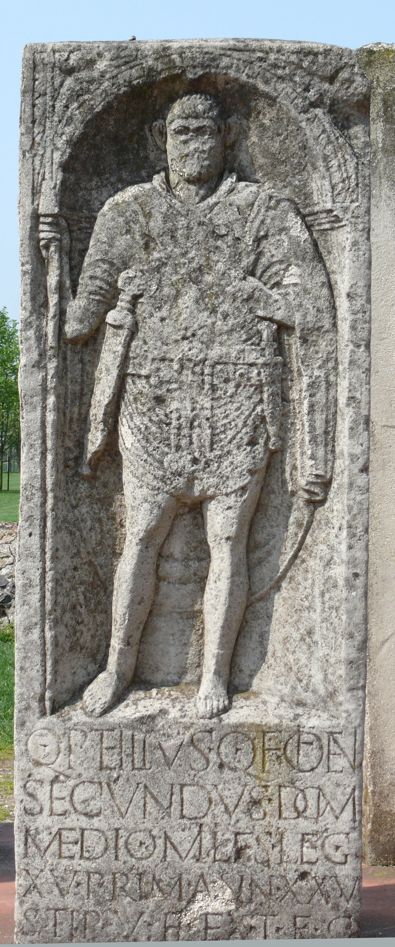 Photo taken in APX, the archeological park in Xanten. For the Latin text, see German version.
Quintus Petilius Secundus, son of Quintus from the tribe of the Oufentina from Milan, soldier of the 15th Legion Primigenia, 25 years old, 5 years in service, the heir has erected this gravestone in compliance with the testament.
The 15th Legion was stationed along with the 5th, in Xanten since 13 AD. Both legions built their camp Vetera in stone. The camp was destroyed by the Bataves during the 69/70 revolt.
Petilius is wearing a tunica and a shoulder cape. He is armed with a lance, a sword and a dagger.